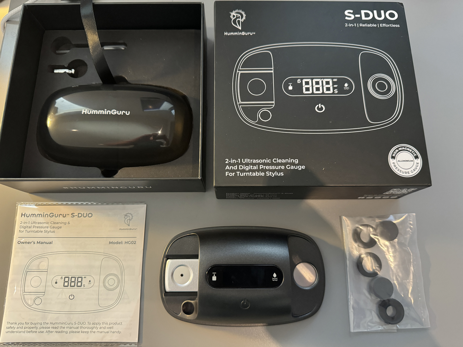 Close-up photo view of HumminGuru S-DUO ultrasonic stylus cleaner/tracking force gauge product model, its product packaging box, its accessories, and its owner's manual nearby