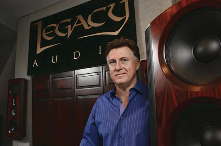 Landscape orientation photograph close-up view of Bill Dudleston, Legacy Audio's Chief Designer, grinning in a dark blue colored button-up dress shirt and standing posing right nearby Legacy Audio product models as well as there happens to be a Legacy Audio banner signage behind him