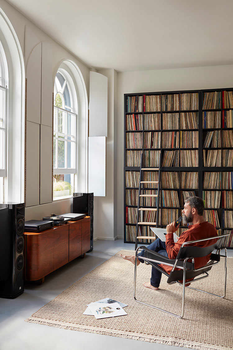 Man seated down on chair in living room area near a bookshelf as he glances out at the window and underneath the window is a audio products setup consisted of an ARCAM integrated amplifier, a turntable, a streamer, and loudspeakers