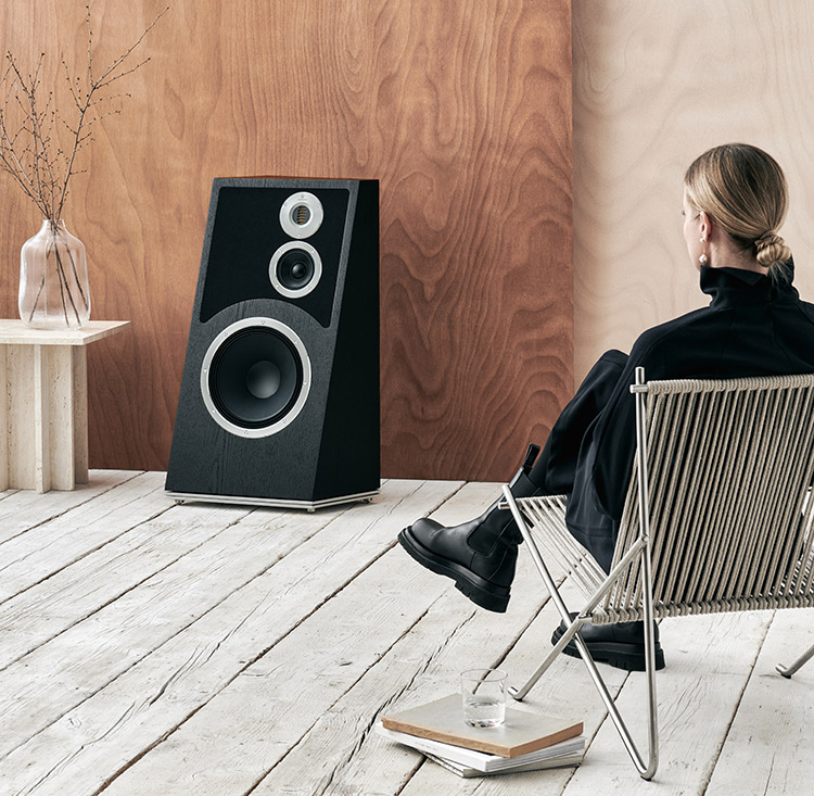 Close-up photograph view of a woman seated down in a chair looking at the Audiovector Trapeze Reimagined 3-way floor standing loudspeaker Living Room Lifestyle View