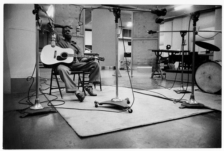 Vintage black and white photograph of John Lee Hooker seated down in a chair holding a guitar as he is in a recording music session room