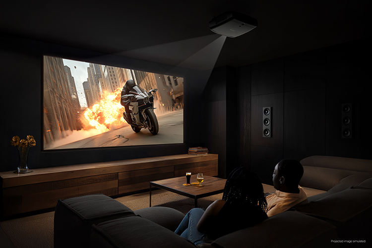 Digital promotional marketing landscape orientation image display of the Epson Pro Cinema LS9000 4K HDR Laser Projector being used in effect in a dimly dark shadow living room area projecting an image of a movie or TV show scene showcasing a person in a motorcycle outfit speeding off in a motorcycle with a fire burst explosion behind him in a cityscape area as this signifies an action drama thriller genre storyline plot with the back rear side view of a woman and man sitting next to each other back on the couch watching this projected image with a small fine print at the right edge corner that says Projected image simulated