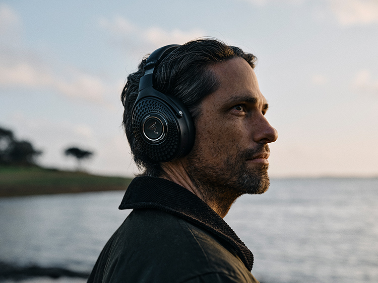 Portrait orientation close-up outdoor photograph view of a man wearing the Focal Bathys Wireless Hi-Fi Headphone Deep Black Finish around his ears as he glances upward at something off into the distance as behind him in the far distant background is a body of water and partial land stretch area filled with trees