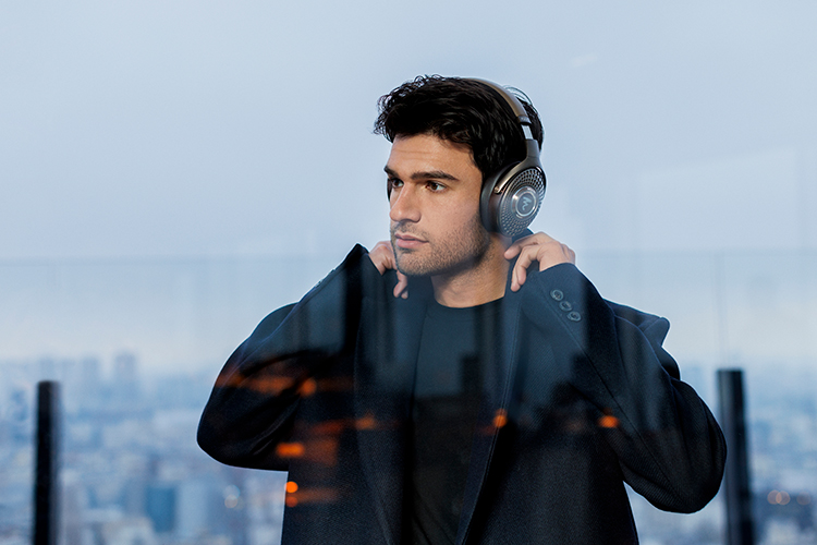 Digital promotional marketing image of a man in a black coat and black sweatshirt underneath lifting up his upper top coat collars as he is listening to his Focal Bathys MG Headphone product model around his ears