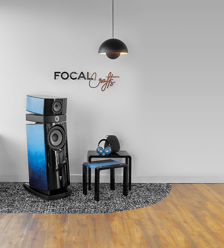 A modern living audio space featuring a single large Focal Utopia Scala EVO 3-way floor-standing speaker with a glossy black and gradient blue color finish; The speaker is on a black and white color patterned rug next to two small, black, square stools; A pair of blue and black Focal headphones is resting on the taller stool; A copper-colored FOCAL CRAFTS logo is on the white wall above the speaker and a single black pendant light hangs from the ceiling