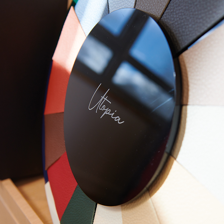 A close-up, angled shot of a round, dark-colored object with the word Utopia engraved in script; This disc is placed over a color wheel showing a variety of leather or fabric swatches in different shades of red, blue, brown, and white