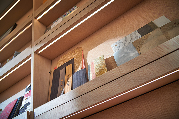 A close-up, angled view of a section of illuminated light wood shelving displaying various material samples for custom design projects; The samples include different types of wood veneer, stone, and other materials; Some pieces are labeled Utopia