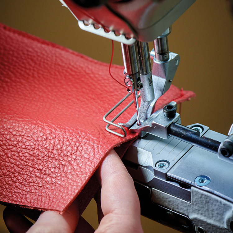 A close-up shot of a sewing machine in operation, with a finger gently guiding a piece of textured red leather under the needle; A red thread is visible in the needle