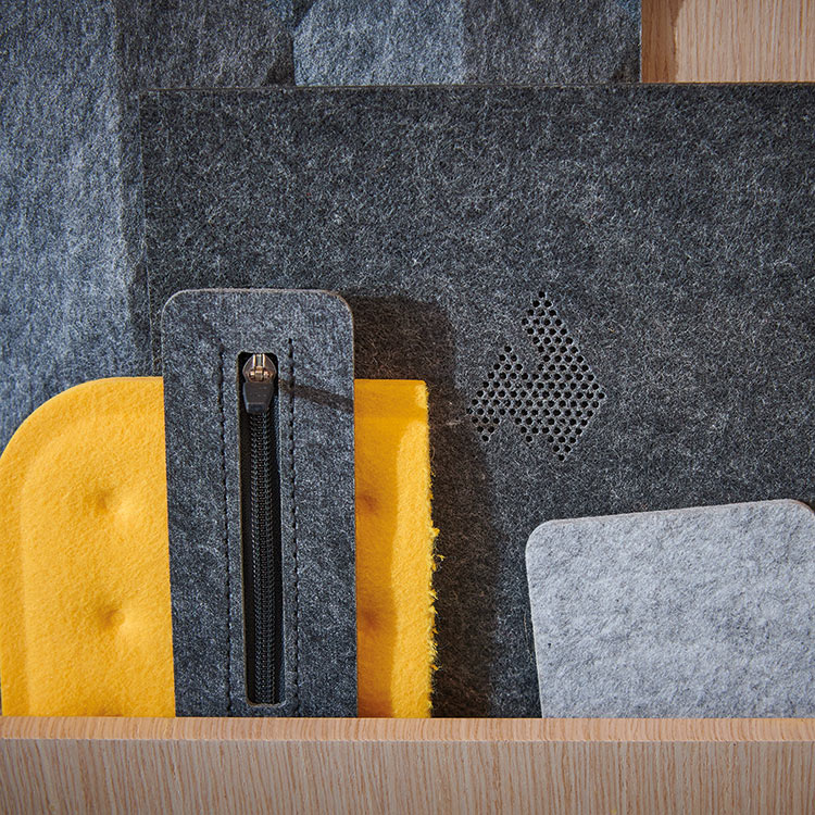 A close-up, high-texture photo of various felt and foam material swatches in shades of dark gray, light gray, and bright yellow; One dark gray piece has a zipper, and another has a hexagonal speaker-like mesh pattern cut into it; The materials are displayed on a light wood shelf