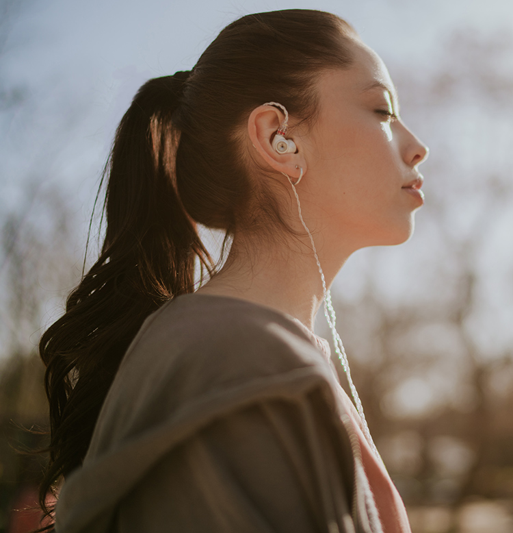 Outdoor portrait photo side view of a woman listening to her Meze Audio ALBA in-ear earphones as the woman has her eyes closed and has a dark forest green jacket on