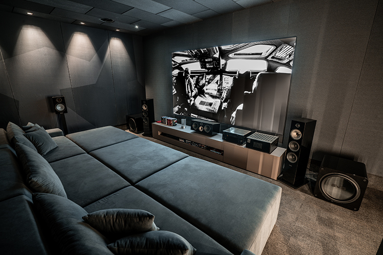 Landscape orientation photograph angle view of a high-end home theater cinema room with dark gray acoustic wall panels and a large, multi-section sofa; A widescreen movie is playing on the what seems to be either a projector or television screen, showing a black and white image scene of some sort; A media cabinet holds several pieces of various assorted home theater high fidelity audio equipment, flanked by tall speakers and a large subwoofer; Spotlights in the ceiling illuminate the area