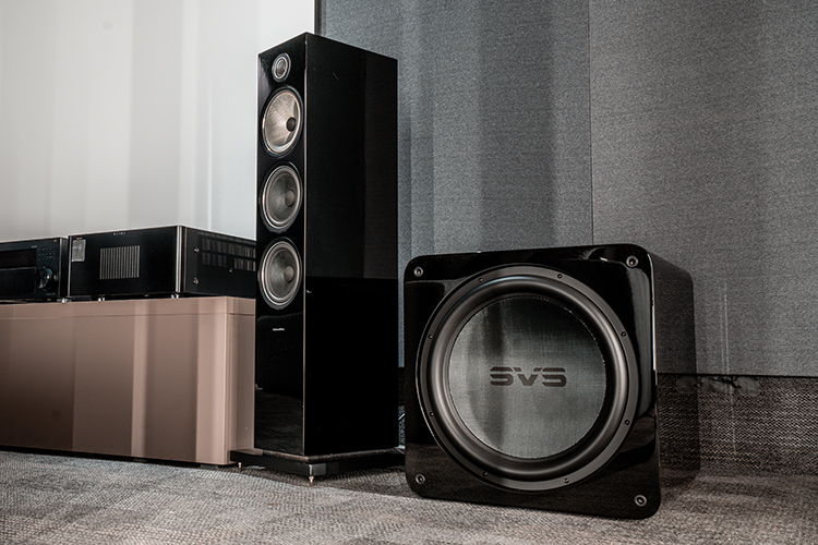Landscape orientation photograph of a high-end home theater cinema room area showcasing a media cabinet that holds several pieces of various assorted home theater high fidelity audio equipment flanked by a tall Bowers and Wilkins speaker and a SVS SB17-Ultra R|Evolution subwoofer