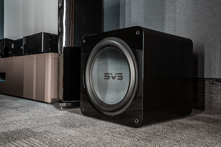 Landscape orientation photograph of a high-end home theater cinema room area showcasing the SVS SB17-Ultra R|Evolution subwoofer nearby a media cabinet that holds several pieces of various assorted home theater high fidelity audio equipment flanked by a tall Bowers and Wilkins speaker