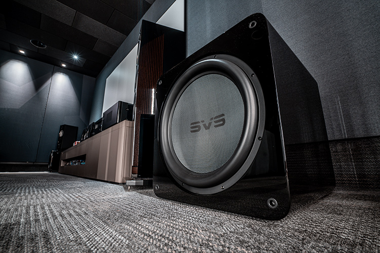 Landscape orientation photograph ground level front angle side view of a high-end home theater cinema room area showcasing the SVS SB17-Ultra R|Evolution subwoofer alongside a media cabinet that holds several pieces of various assorted home theater high fidelity audio equipment flanked by a tall Bowers and Wilkins speaker; Spotlights in the ceiling illuminate the area