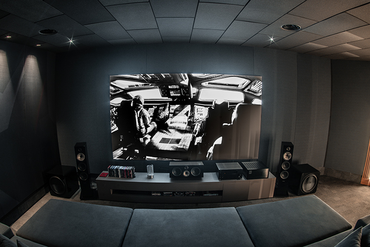 Landscape orientation photograph angle view of a high-end home theater cinema room with dark gray acoustic wall panels and a large, multi-section sofa; A widescreen movie is playing on the what seems to be either a projector or television screen, showing a black and white image scene of some sort; A media cabinet holds several pieces of various assorted home theater high fidelity audio equipment, flanked by tall speakers and a large subwoofer; Spotlights in the ceiling illuminate the area