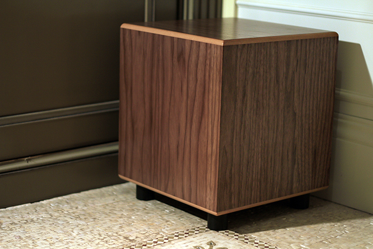 MJ Acoustics Reference 100 MkII Subwoofer American Black Walnut Finish Angle View sitting on short black feet on a tiled floor; It is positioned near a doorway and a white wall corner