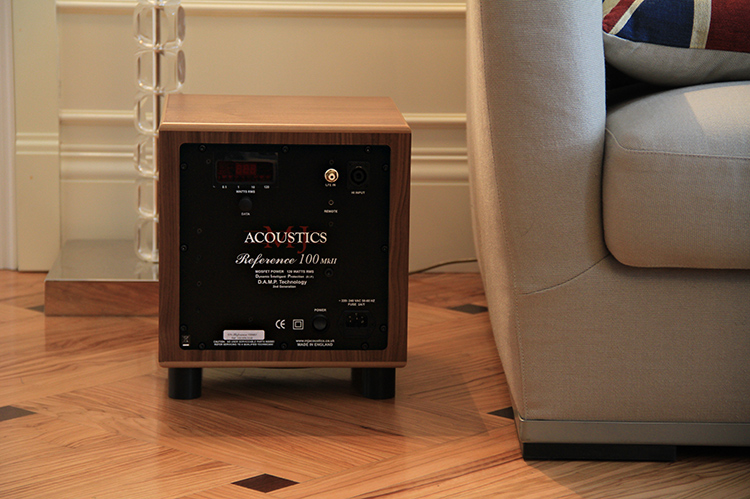 MJ Acoustics Reference 100 MkII Subwoofer American Black Walnut Finish Rear View sitting on short black feet on a tiled floor; It is positioned near a beige colored couch and partial white area of a wall border