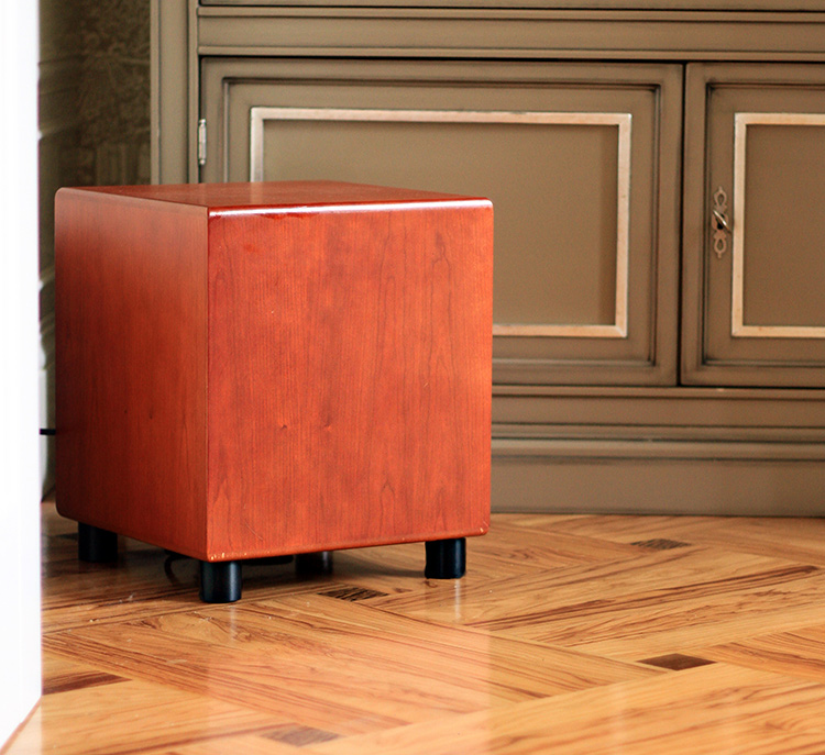 MJ Acoustics Reference 200 Subwoofer Cherry Finish Angle View sitting on short black feet on a tiled floor; It is positioned near a dark grey drawer cabinet and partial white area of a wall border