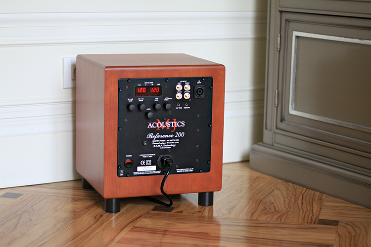 MJ Acoustics Reference 200 Subwoofer Cherry Finish Rear View sitting on short black feet on a tiled floor; It is positioned near a dark grey drawer cabinet and partial white area of a wall border