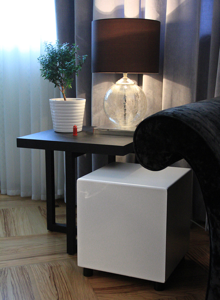 MJ Acoustics Reference 400 MkI Subwoofer White Satin Finish Angle View sitting on short black feet on a tiled floor; It is positioned near a dark navy blue couch chair and underneath a black tabletop stand that contains a planter and a lamp