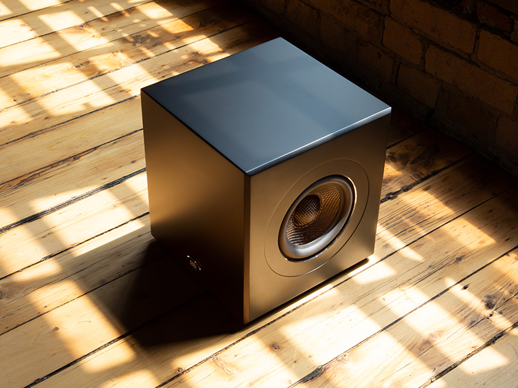 An overhead, angled shot of a compact, matte black PSB Speakers SubSeries BP7 Powered Subwoofer with a single exposed circular driver on the front panel; The subwoofer is sitting on a wooden floor with a window pane shadow casting diagonal lines of light across the floor; A brick wall is visible in the background