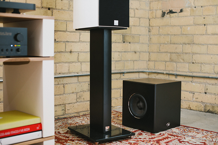 A compact, black PSB Speakers SubSeries BP7 Powered Subwoofer is sitting on a patterned rug next to a tall, black DALI speaker stand holding a white smaller DALI bookshelf speaker with a white and black cabinet; A brick wall is in the background and a shelving unit with home theater high fidelity audio components and books is partially visible on the left