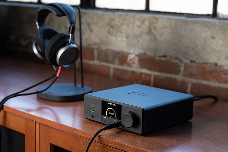Rotel DX-3 High Performance Headphone Amplifier, Pre-Amplifier and DAC Black Finish Angle View situated next to nearby a random headphone product model located on top of a headphone stand display as everything is on a wooden table