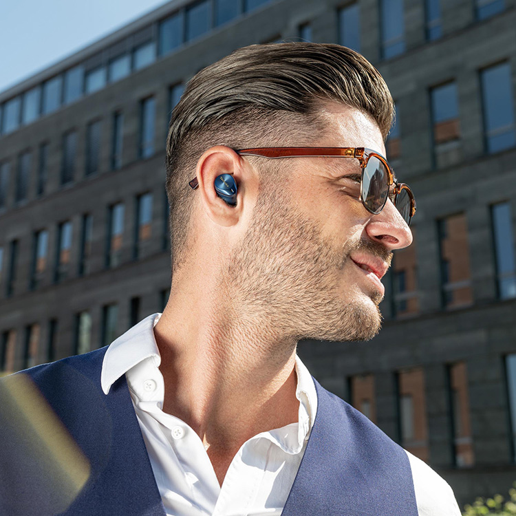 Man in sunglasses and business suit blazer standing outside nearby a building with numerous windows while he is smiling as he listens to his Technics EAH-AZ80 True Wireless Earbuds Midnight Blue Finish