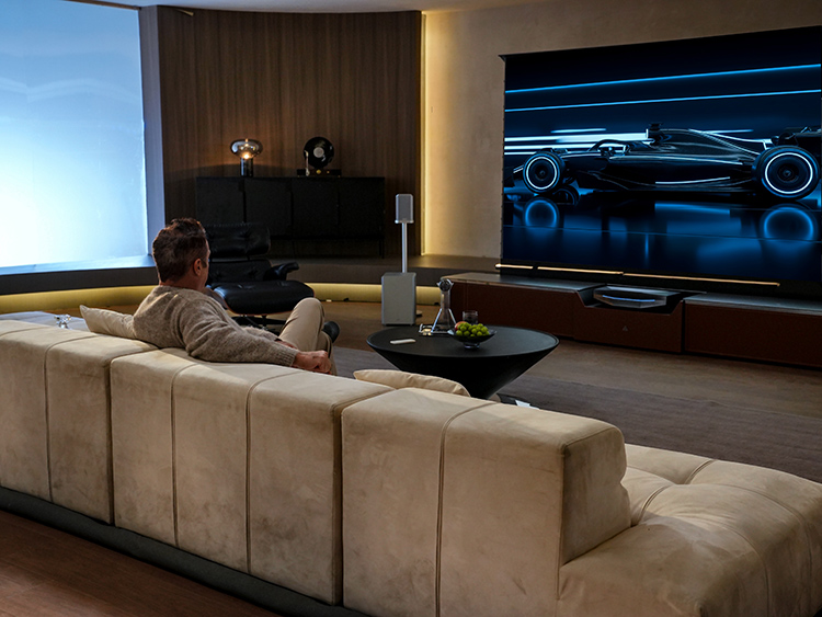 Male person seated in a living room couch watching television with the The Grand Vanish TV Masterpiece Bundle featured which includes the AWOL Vision Aetherion Max Projector, 130
