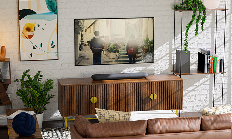 Digital image display landscape orientation view of a modern living room with a TV mounted above a fluted wood media console with yellow circular handles; A cylindrical Bluesound PULSE CINEMA – Black Soundbar is sitting on top of the console; The TV screen shows two children walking up an outdoor staircase; The living room has a white brick wall, a brown leather couch, and various plants and decor