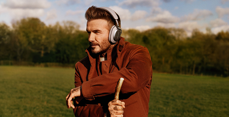 A landscape orientation photograph marketing promotional image display of David Beckham, a man with dark brown hair, a dark brown beard, and a thick dark reddish-brown jacket is standing in a grassy field amongst nature; He is wearing the Bowers and Wilkins Px8 S2 Warm Stone color finish over-ear wireless headphones and holding a wooden walking stick; He is looking to his left, away from the camera