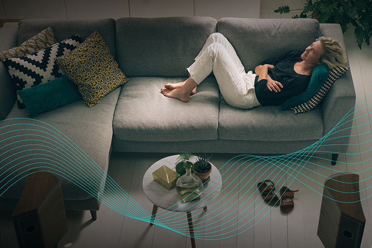 A woman is laying down on the couch in the living room listening to her home theater hifi sound devices with the Dirac Live Active Room Treatment function active displayed in blue curvy graph line angles