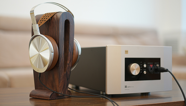 HIFIMAN SUSVARA UNVEILED Reference Open-Back Planar Headphone resting on top of a wooden headphone pedestal stand connected by cables to a HIFIMAN amplifier product model