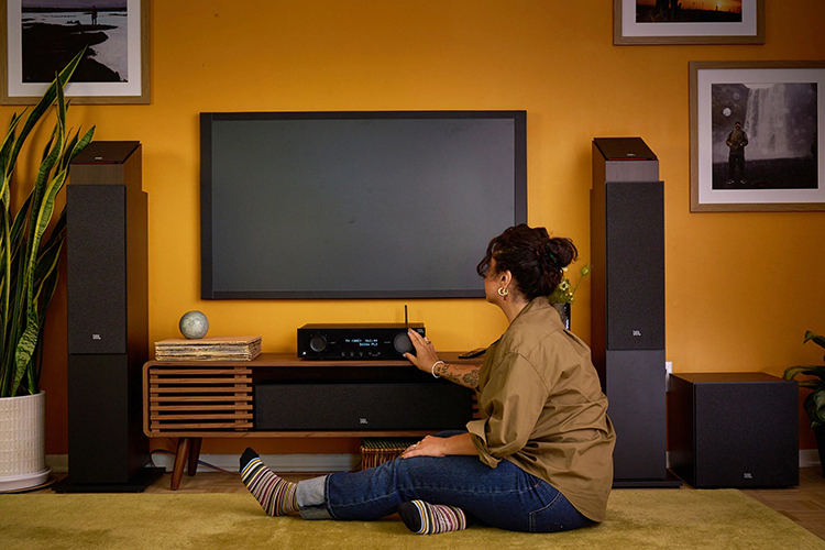 Living room lifestyle close-up landscape photograph view of a woman seated down as she is turning the dial button with her hand on the JBL Modern Audio (MA) AV Receiver product model, which is located underneath the flat screen wall tv plus there are other JBL product models nearby this woman such as the JBL Stage 2 loudspeakers