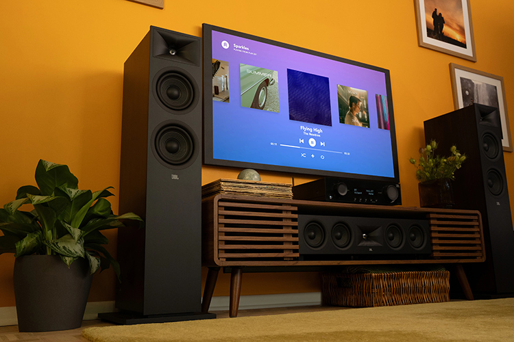 Living room lifestyle close-up landscape photograph angle view of a JBL Modern Audio (MA) AV Receiver product model, which is located underneath the flat screen wall tv plus there are other JBL product models nearby such as the JBL Stage 2 loudspeakers and a Center Channel speaker product model