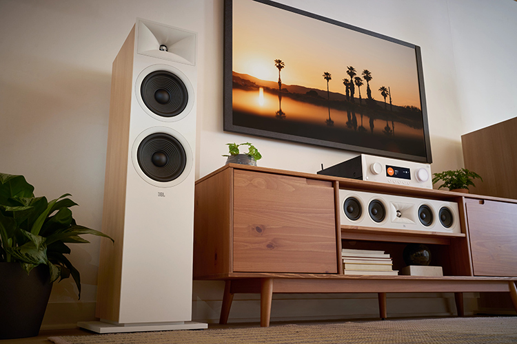 Living room lifestyle close-up landscape photograph angle view of a JBL Modern Audio (MA) AV Receiver product model, which is located underneath the flat screen wall tv plus there are other JBL product models nearby such as the JBL Stage 2 loudspeakers and a Center Channel speaker product model