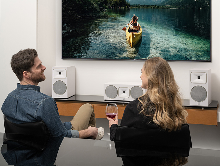 A man and a woman are sitting right next to each other chatting as they look each other in the eyes with the MartinLogan Motion Foundation B2 Bookshelf Speakers (Satin White Finish) and Motion Foundation C1 Center Channel Speaker (Satin White Finish) situated across from them underneath the living room television