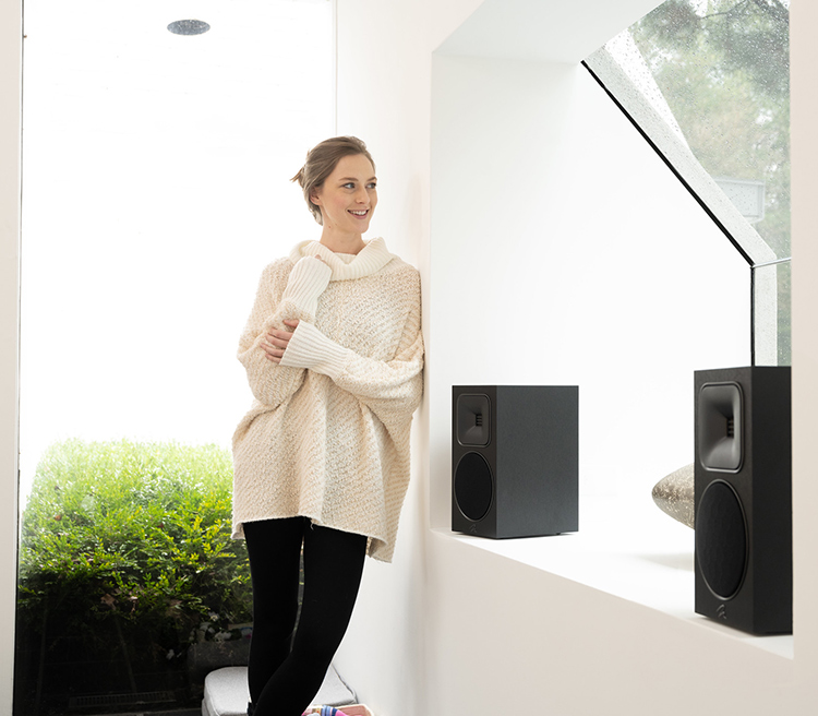 A woman smiles leaning on the side of a wall glancing at the reflection of a window with the MartinLogan Motion Foundation B1 Bookshelf Loudspeakers (Black Finish) situated right next to her
