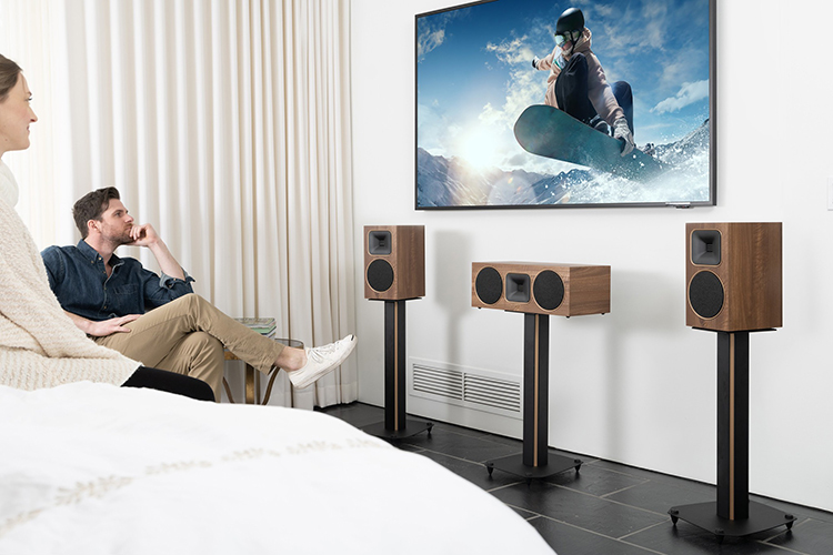 A mand and a woman are glancing at the living room television screen grinning as the MartinLogan Motion Foundation C1 Center Channel Speaker (Walnut Finish) and Motion Foundation B1 Bookshelf Loudspeakers (Walnut Finish) are on a stand situated underneath the living room television screen