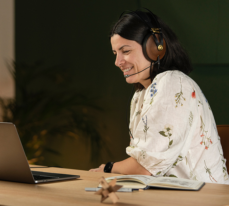 Woman smiling as she has on the Meze Audio 99 Classics Walnut Gold Headphone with the Boom Mic while she stares at her laptop screen with her seated down at a table