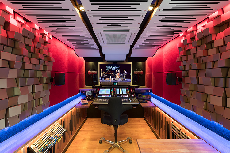 A wide-angle interior view of a highly professional mobile audio mixing studio; The room features vibrant red padded walls and intricate wood-block acoustic diffusers on the sides, with a large digital mixing console in the center facing a screen displaying the Ystad Sweden Jazz Festival