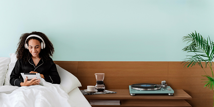TEAC TN 400BT X/TB Turquoise Blue Limited Edition Analog Turntable situated on a wooden desk tabletop nearby a woman resting in bed with her headphones and tablet device