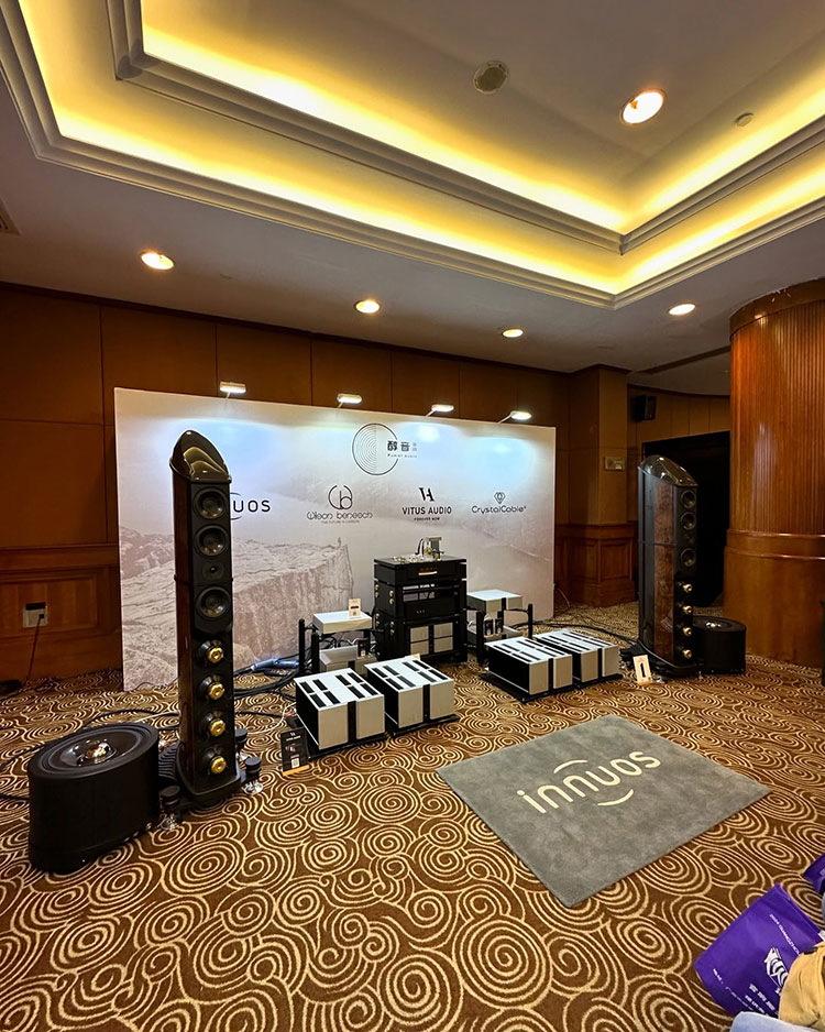 Portrait orientation indoor photograph of various home theater high fidelity audio product models on display within a ballroom showroom area with a Innuous audio brand company logo mat situated in front of these products while behind these products in the background is a banner display featuring different various audio company logos