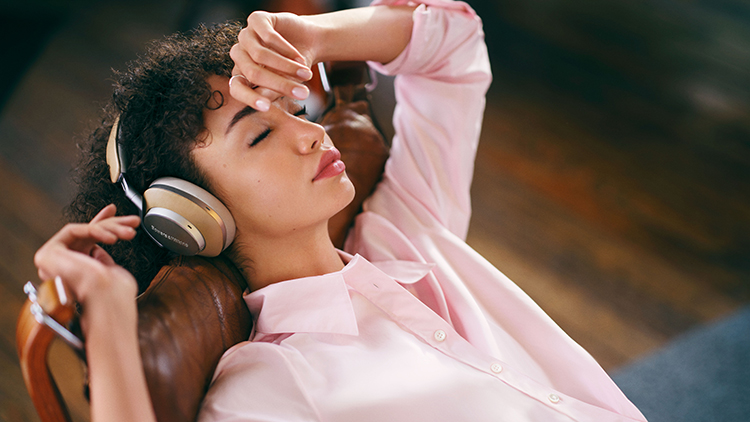 A woman listens with the Bowers and Wilkins Px8 Wireless Headphone Tan Finish on top of her head laying down on a chair