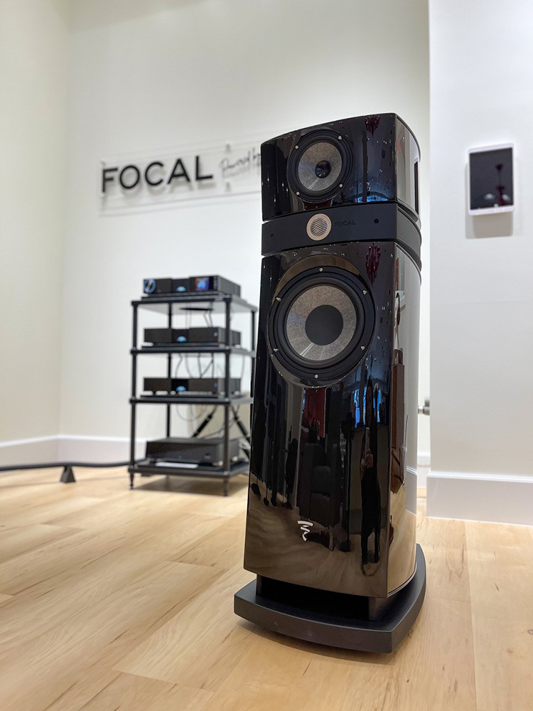 Close-up view of a Focal Powered by Naim floorstanding loudspeaker product in the foreground and other assorted products behind in the background