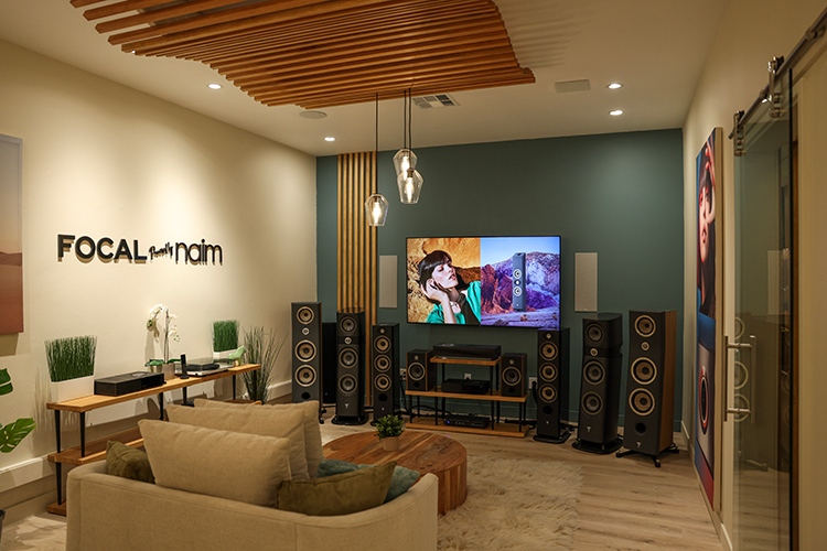 An interior landscape photograph of the Focal Powered by Naim retail space in Newport Beach, California featuring Focal Powered by Naim turntables, loudspeakers, in-wall speakers, subwoofers, and amplifiers in the showroom area located nearby a small sofa chair and a small round circular table