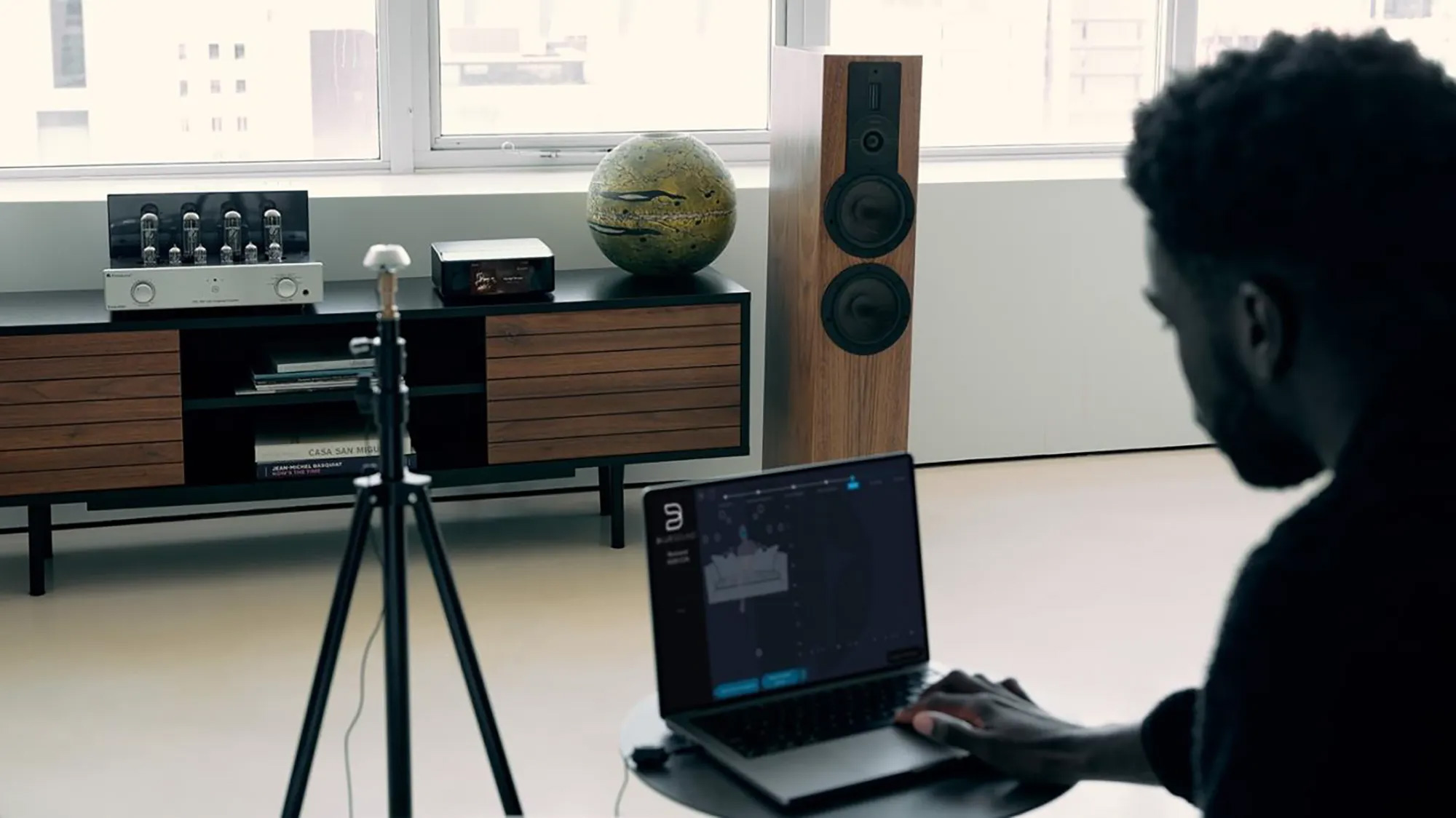 A man operates a laptop running Dirac Live software in a modern living room setup, featuring a wooden floor-standing speaker, an amplifier, and a decorative globe on a media console.