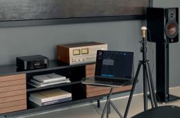 A laptop displaying Dirac Live software is positioned on a small table, connected to audio equipment on a modern media console with a wooden speaker on a stand in the background.