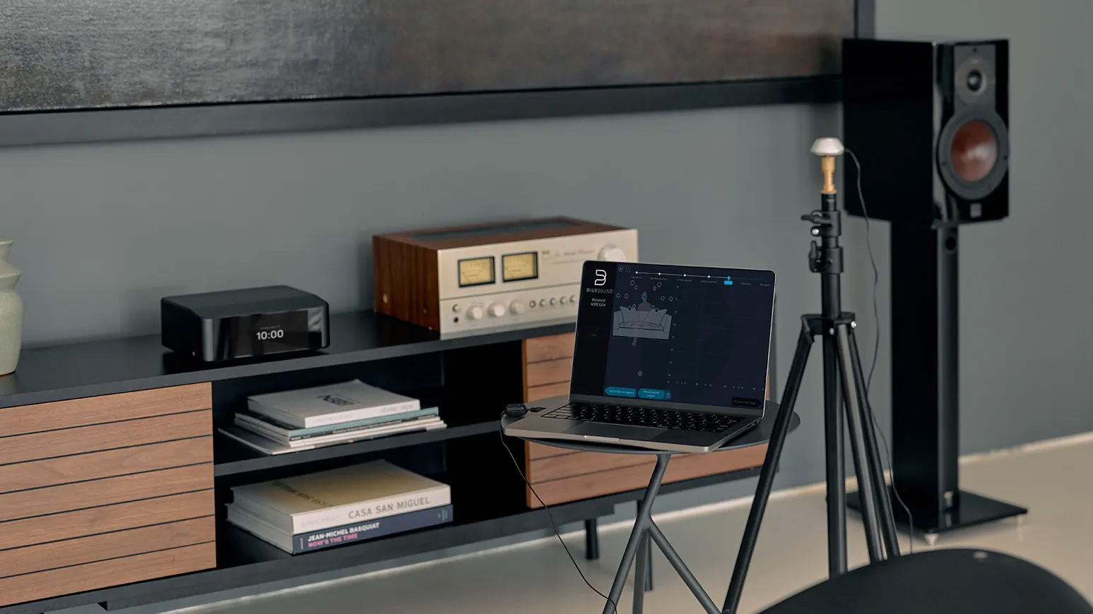 A laptop displaying Dirac Live software is positioned on a small table, connected to audio equipment on a modern media console with a wooden speaker on a stand in the background.